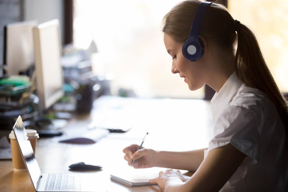 garota escrevendo em bloco de notas com fones de ouvido e a frente de laptop